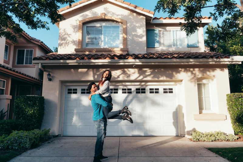 Joyful couple celebrating with raised arms in front of their new home on a sunny day.