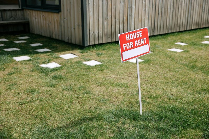 Red 'House for Rent' sign on grassy lawn next to a wooden house exterior.