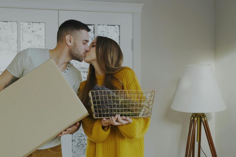 A couple shares a kiss while holding a moving box and decorative basket in their new home.