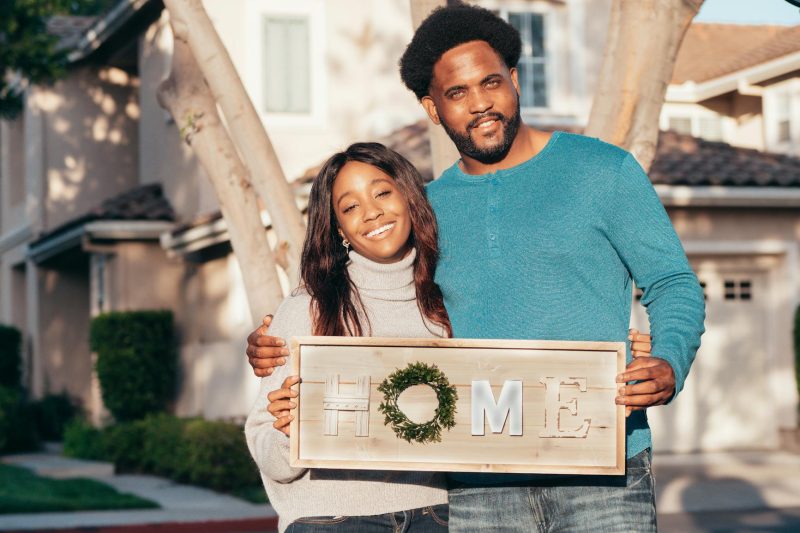 Happy couple holding a 'Home' sign in front of their new house, expressing joy and togetherness.