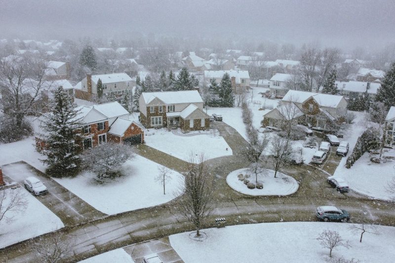 Aerial view of a snowy suburban neighborhood with houses, streets, and trees under a clear sky.