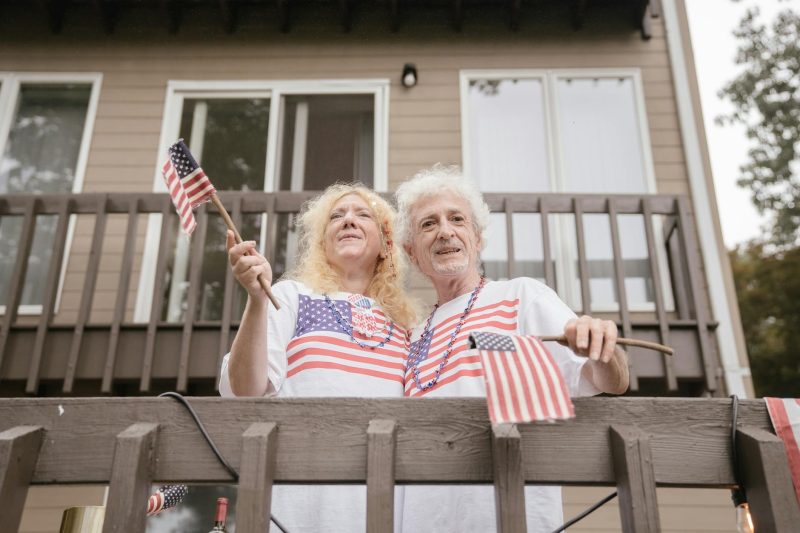 Senior couple on a balcony, smiling and celebrating with US flags in a sunny outdoor setting.