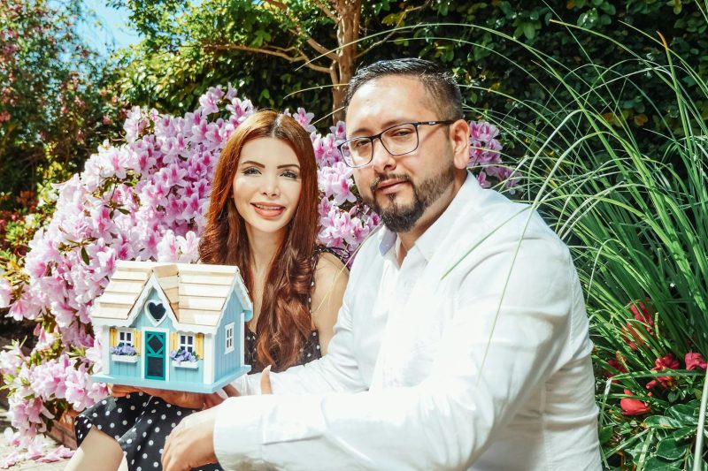 Couple smiling while holding a miniature house, surrounded by colorful garden flowers.