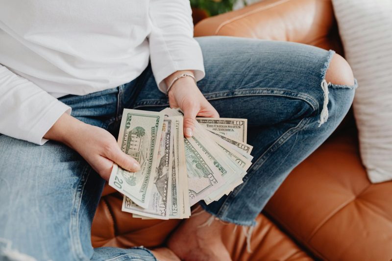 A woman in jeans counts US dollar bills while seated on a leather sofa.
