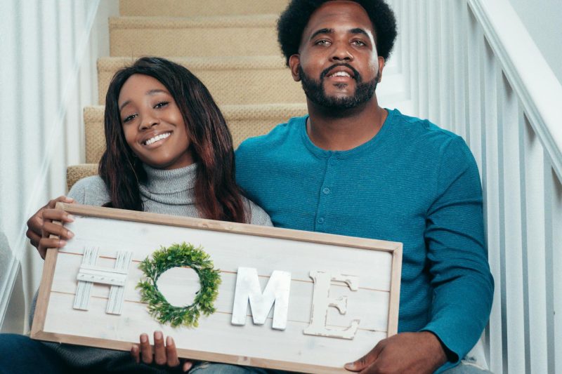 Smiling couple standing on stairs, holding a decorative 'Home' sign, celebrating new home ownership.