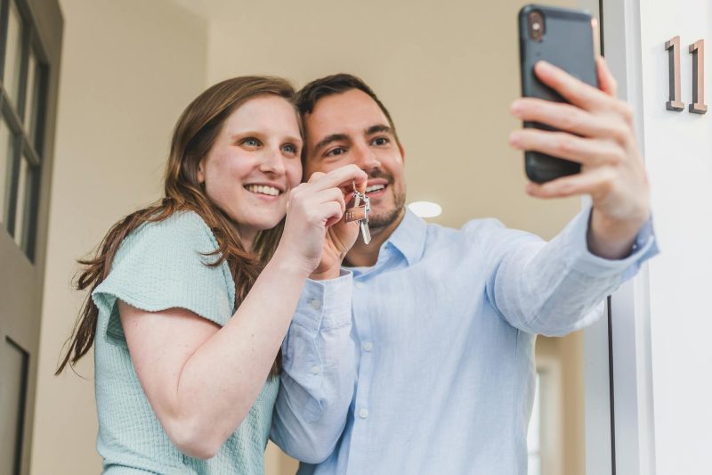 Joyful couple smiling and holding keys to their new home while taking a selfie.